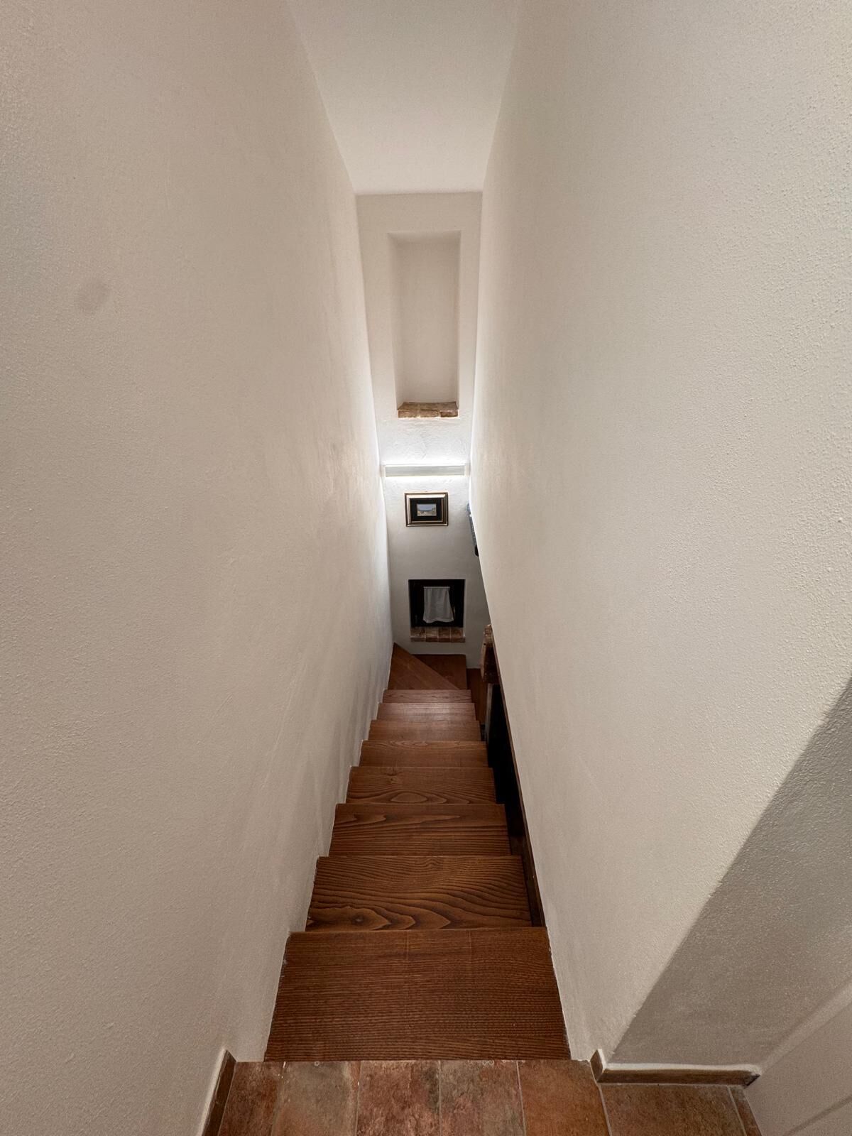 Eine enge Treppe mit Holzdielen und weißen Wänden, die zu einer Empfangshalle mit einem gerahmten Bild und einem kleinen Fenster führt.