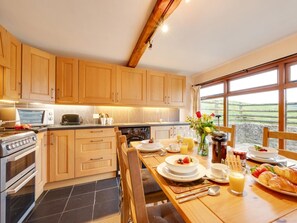 A kitchen with wooden cabinets, a stove, a microwave, and a dining area with a table set for breakfast. - 2 Bed in South Molton (oc-88992) (Bishops Nympton)