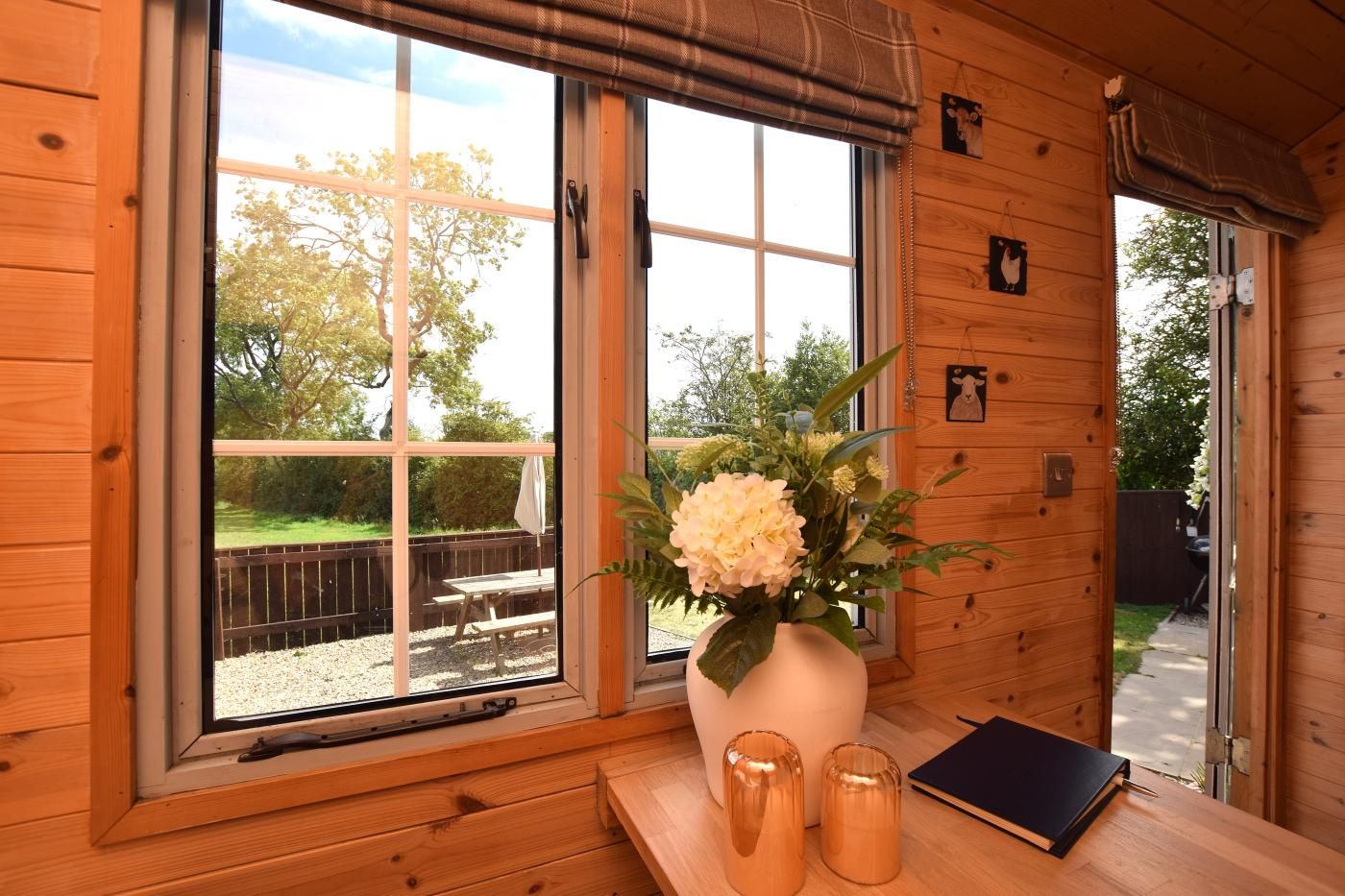 A wooden cabin interior with a large window, a wooden bench, a vase with flowers, and a small table with a notebook.