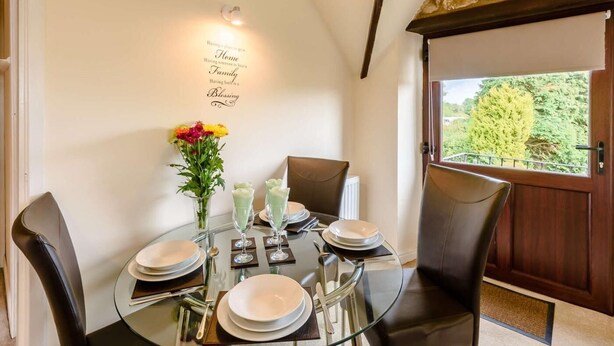 A dining area with a round glass table set for four, a stone wall, and a view of greenery outside. - 2 Bed in Pickering (oc-88957) (Newton-on-Rawcliffe)