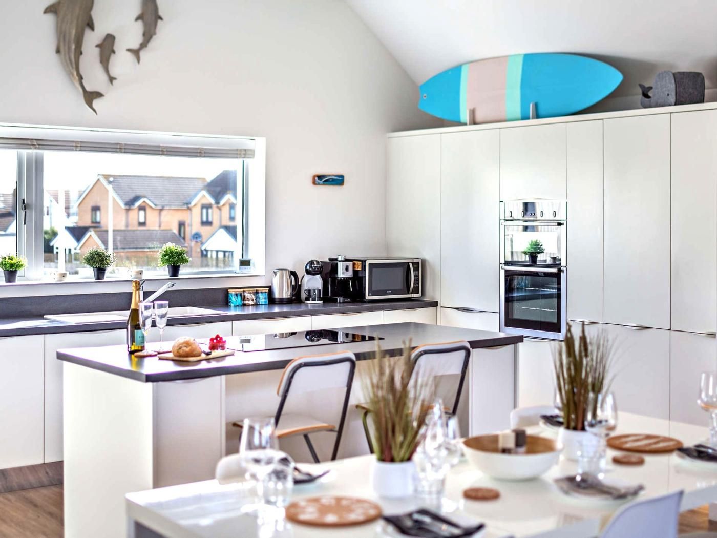 A modern kitchen with a central island, a dining area with a table set for two, and a view of houses through the window.