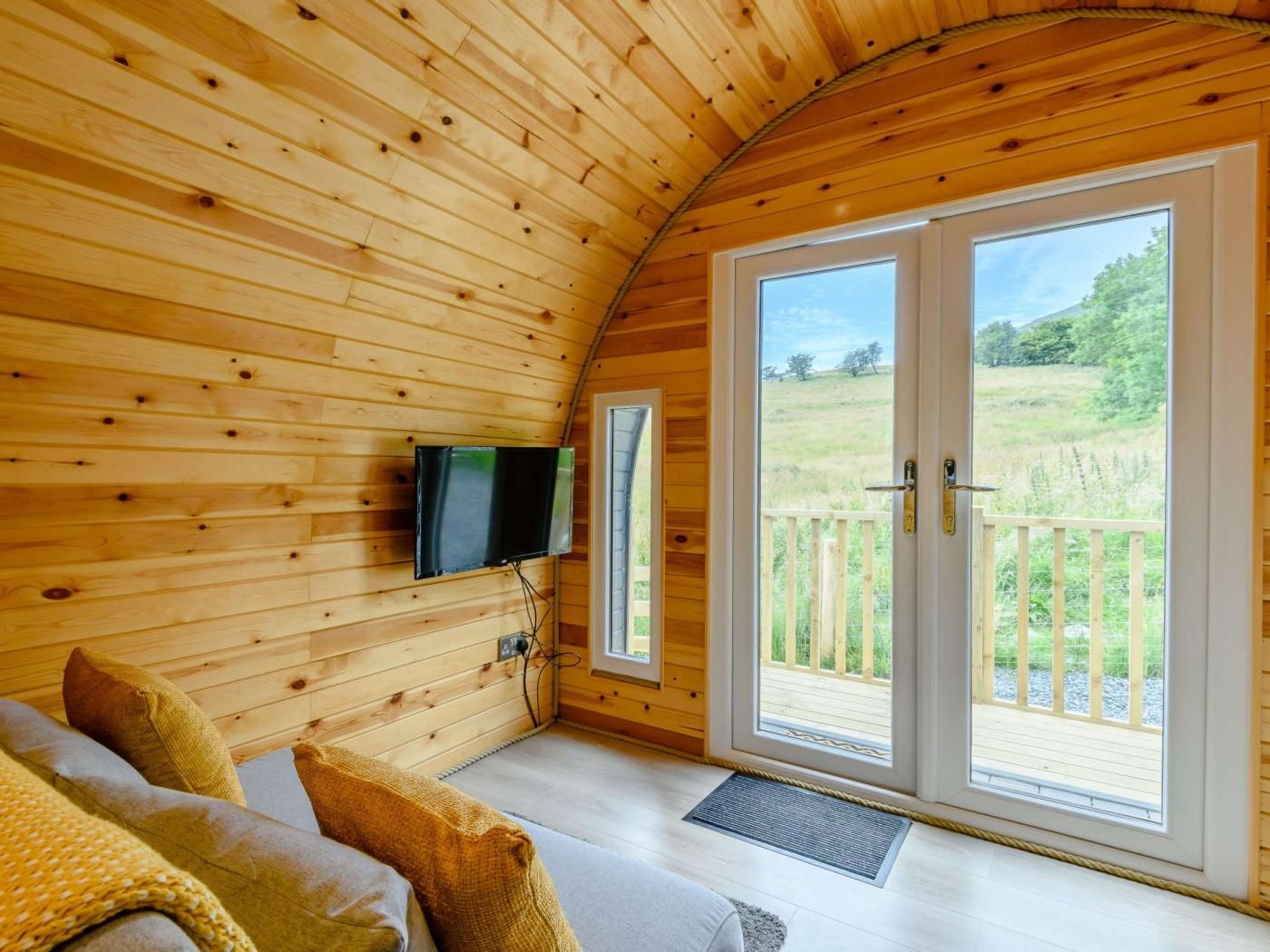 L’intérieur chaleureux d’un chalet avec un plafond en bois, un téléviseur à écran plat fixé au mur, un canapé avec des coussins et une grande porte-fenêtre coulissante donnant sur une terrasse extérieure.