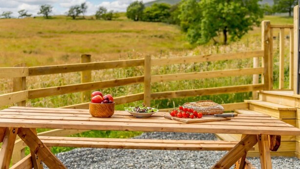 A wooden picnic table with a bowl of fruit, a plate of salad, and a loaf of bread.