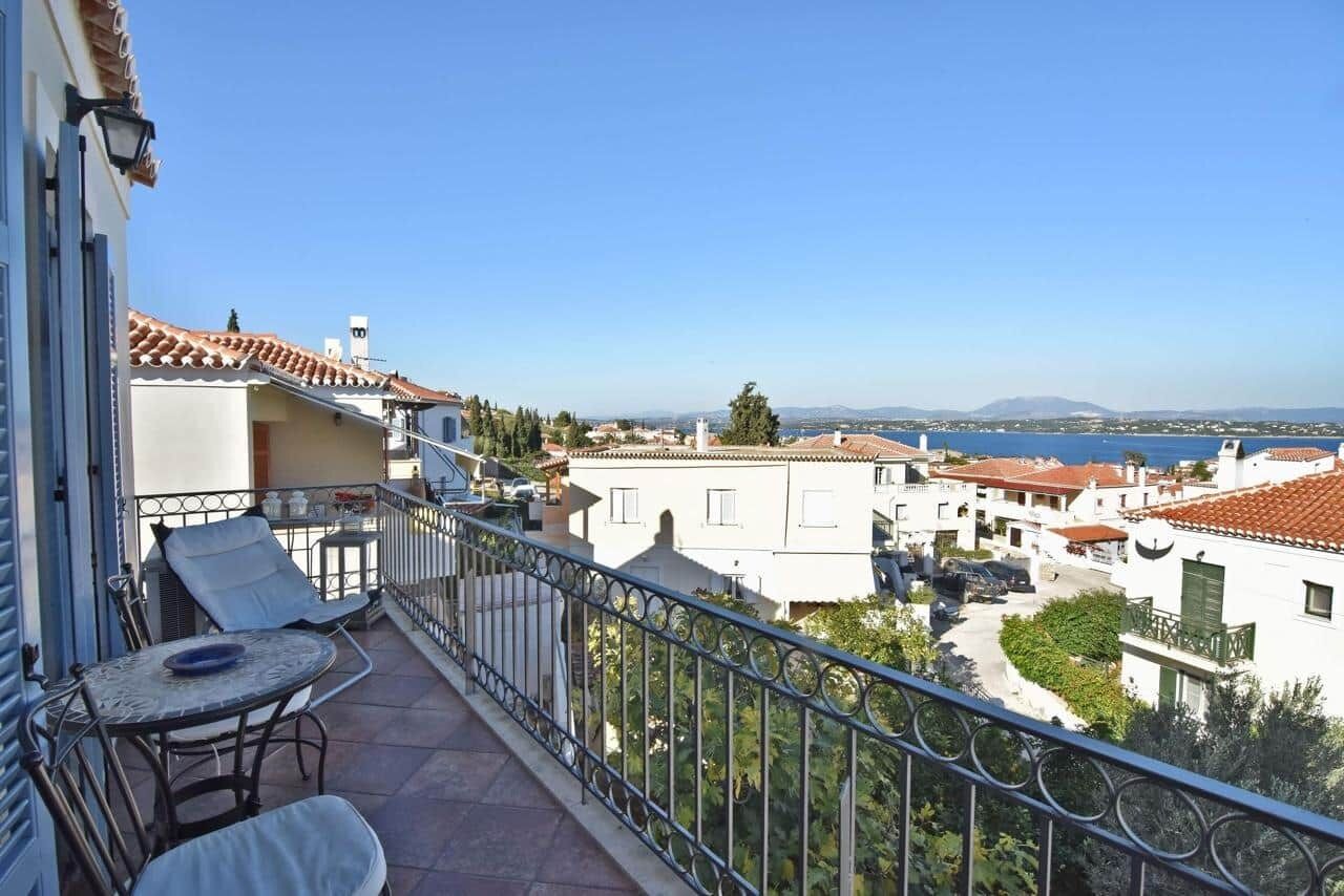 Un balcon avec une table et des chaises, donnant sur une ville côtière avec ses bâtiments et un ciel dégagé.