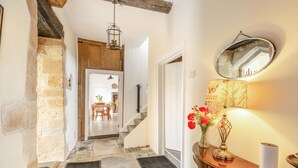 Un couloir avec un sol en pierre, un escalier en bois, un coin repas à l’arrière-plan, un miroir et une lampe décorée de fleurs.