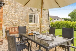 An outdoor dining area with a stone wall, a wooden bench, and a table set for tea service. - 3 Bed in Bridport (oc-89004) (Wootton Fitzpaine)
