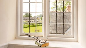 Une fenêtre donnant sur un jardin et des bâtiments, une paire de lunettes et un livre posés sur le rebord de la fenêtre.