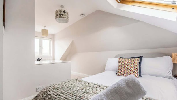 Une chambre avec un plafond en pente, un lit avec une literie blanche, un coussin coloré, une fenêtre et un luminaire fixé au mur.