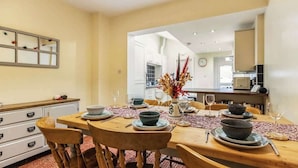 Un coin repas avec une table en bois dressée pour quatre personnes, entourée de chaises. Un vase contenant des fleurs séchées est posé sur la table, et une horloge est accrochée au mur.