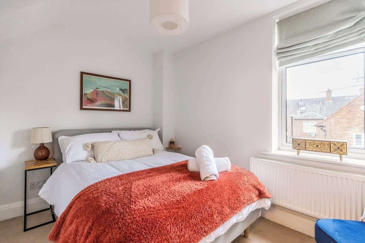 Ein Schlafzimmer mit Bett, Nachttisch, Lampe, einem Gemälde, einem Fenster mit Blick auf Gebäude und einem blauen Sessel.