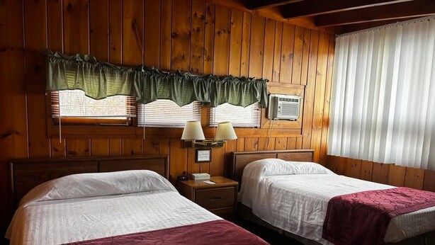 Una habitación con dos camas, paredes de madera, un ventanal con persianas y una mesita de noche con lámparas.