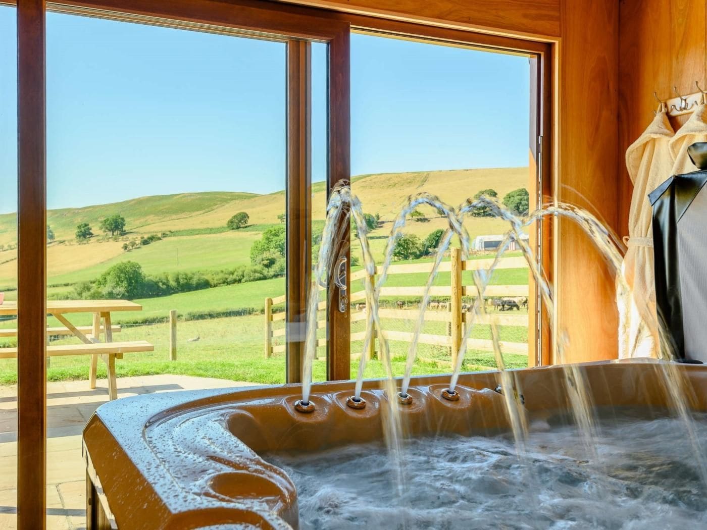 A hot tub with multiple faucets, a wooden bench, and a view of a green landscape through large windows.