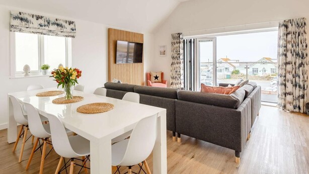 Een moderne woonkamer met een bank, een eettafel met stoelen, een flatscreen televisie bevestigd aan een houten paneel en grote ramen met gordijnen.