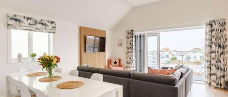A modern living room with a sofa, a dining table with chairs, a flat-screen TV mounted on a wooden panel, and large windows with curtains.