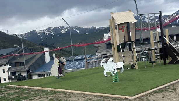 Ein Spielplatz mit einer Kletteranlage, einem kleinen Gebäude und einer Bergkulisse.