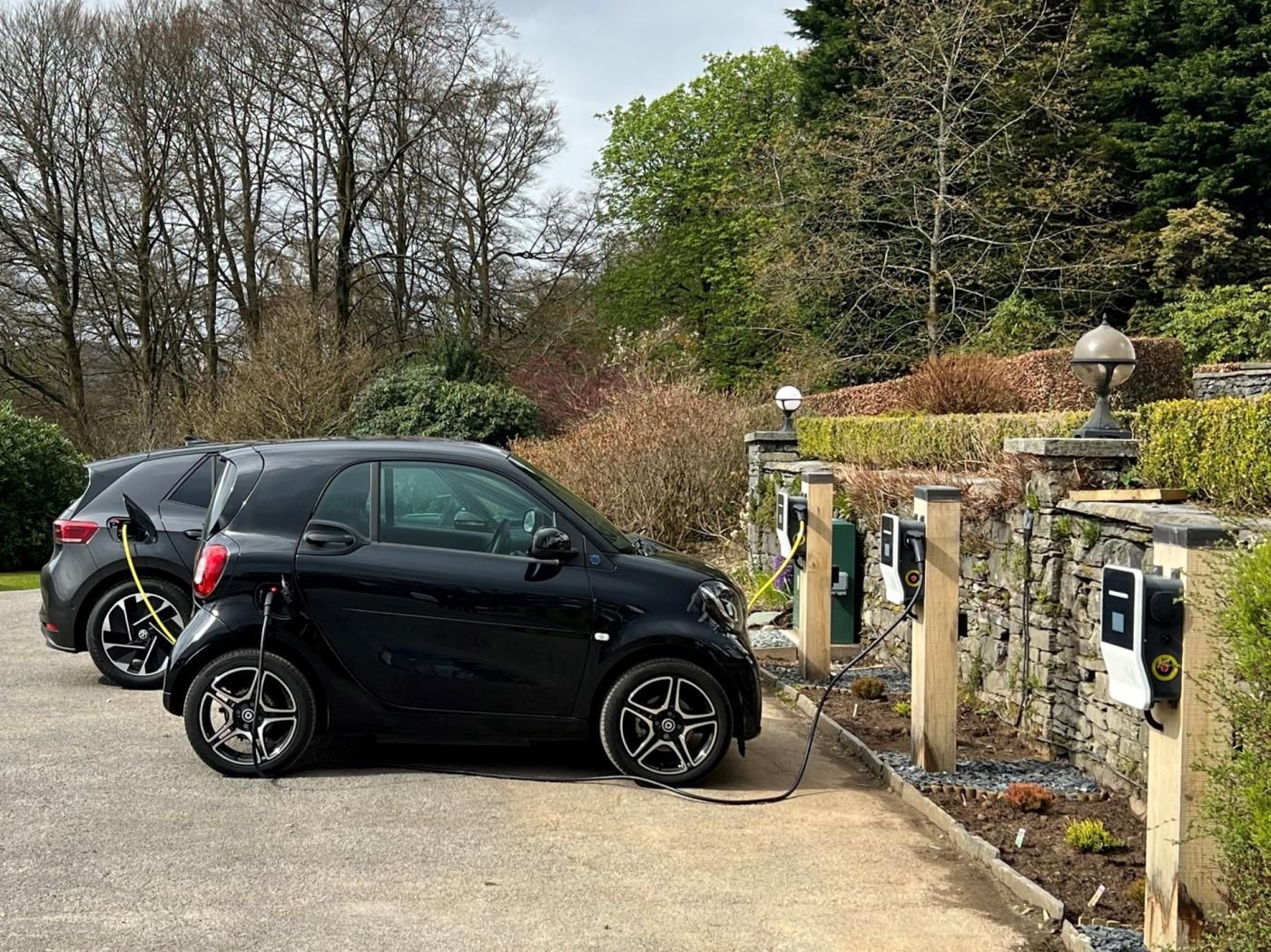 A black car parked at an electric charging station with a stone wall and greenery in the background.