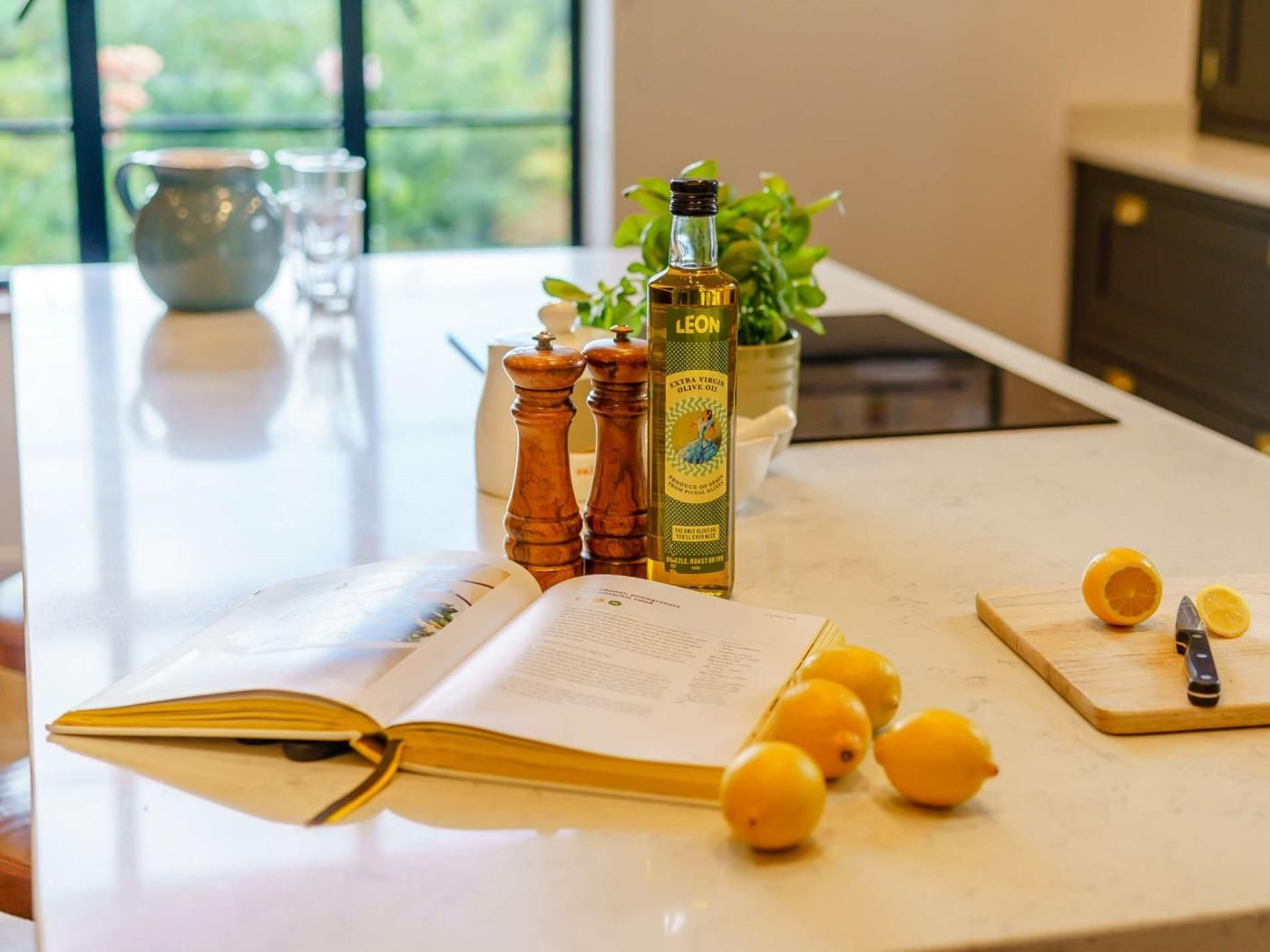 Un plan de travail de cuisine avec un livre, des citrons, une bouteille d’huile d’olive extra vierge et une planche à découper avec un couteau.