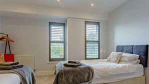 A bedroom with two beds, a radiator, a window with blinds, a bedside lamp, and a hanging coat rack. - 2 Bed in Sandown (oc-89291) (Sandown)