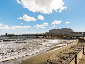 A beach with a pier extending into the water, a building labeled 'Sandcastle Inn,' and a clear sky with clouds. - 2 Bed in Sandown (oc-89291) (Sandown)