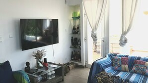 Een woonkamer met een flatscreen televisie aan de muur, een bank met decoratieve kussens, een bijzettafeltje met decoratieve objecten en een boekenkast met diverse spullen.