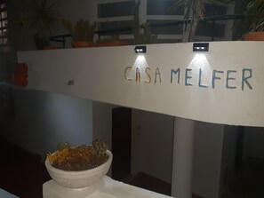 Eine weiße Wand mit einem Schild, auf dem 'CISA MELFER' steht, und einem Blumentopf mit gelben Blüten.
