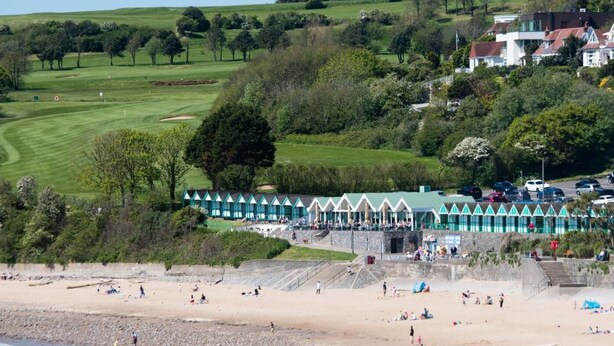 Eine Küstenlandschaft mit Strand, einer Reihe Strandhäuschen und einem Golfplatz im Hintergrund.