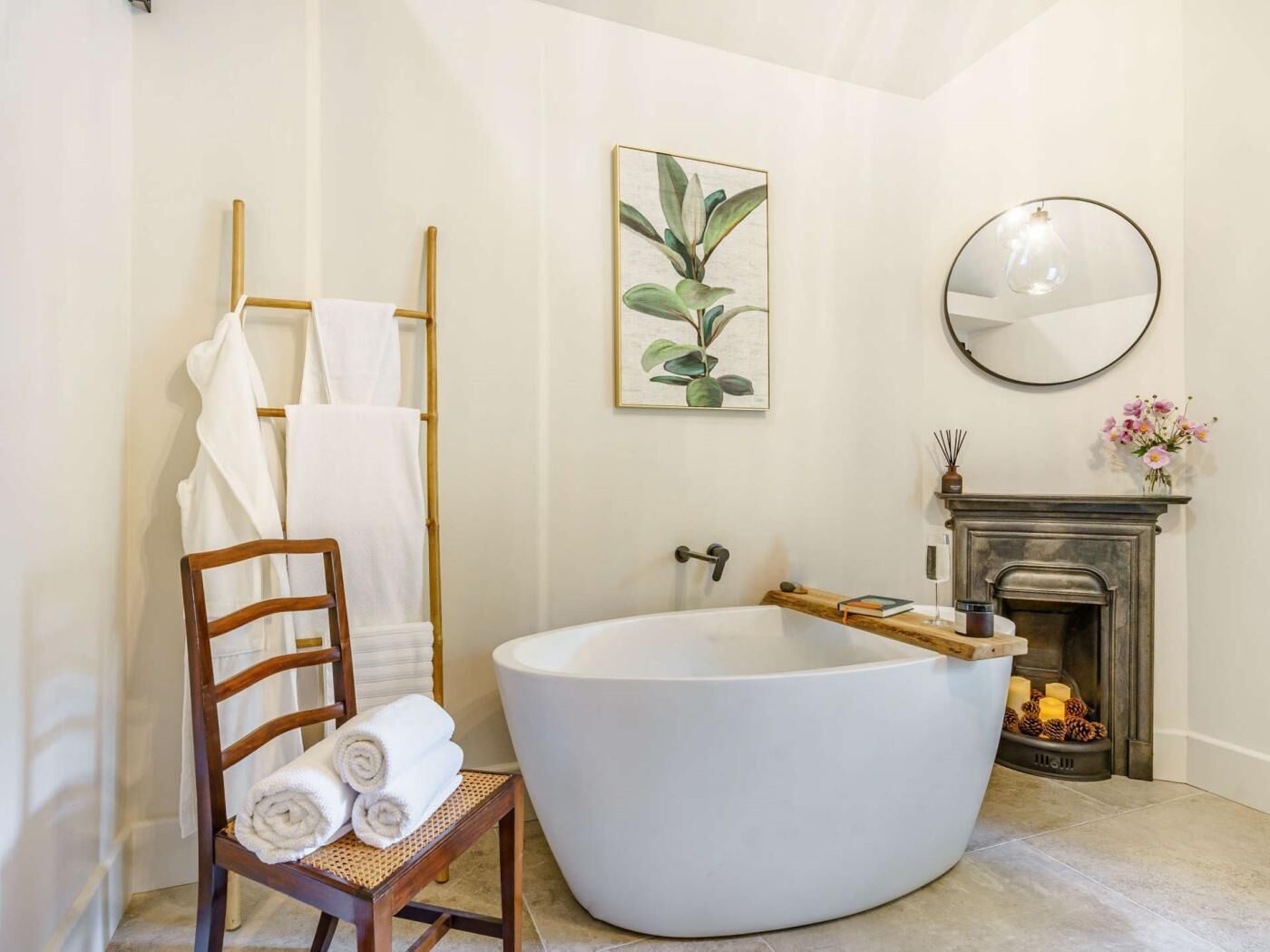 A bathroom with a freestanding bathtub, a wooden chair with towels, a framed plant, a round mirror, and a fireplace with decorative items.