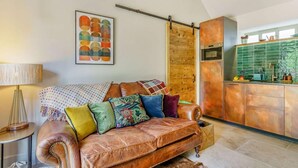 A cozy living room with a leather sofa, a patterned throw pillow, a lamp, and a kitchen area with a green backsplash and copper appliances. - 1 Bed in Durham (oc-89318) (Witton Gilbert)