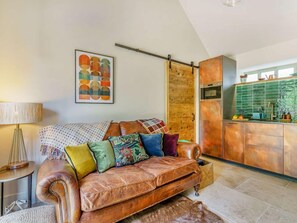 Ein gemütliches Wohnzimmer mit einem Ledersofa, einem gemusterten Zierkissen, einer Lampe und einer Küchenzeile mit grüner Spritzschutzleiste und Kupfergeräten.