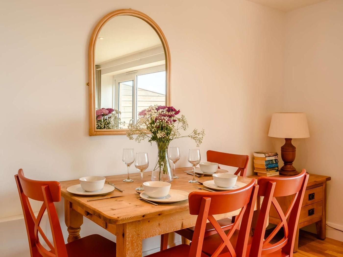 A dining area with a wooden table set for four, a vase of flowers, and a mirror above the table.