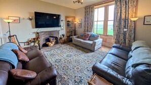 A living room with leather sofas, a fireplace, a television, and a large window with curtains. - 5 Bed in Melrose (oc-89348) (Newtown Saint Boswells)