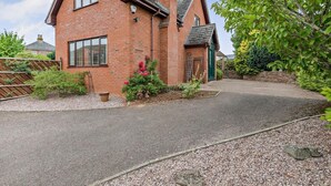 A red brick house with a sloped roof, a front door, and a driveway leading to the entrance. - 3 Bed in Ross-on-wye (oc-89313) (Bridstow)