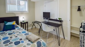 Ein Schlafzimmer mit Bett, Schreibtisch, Stuhl, Fernseher und einem Fenster mit Jalousien.