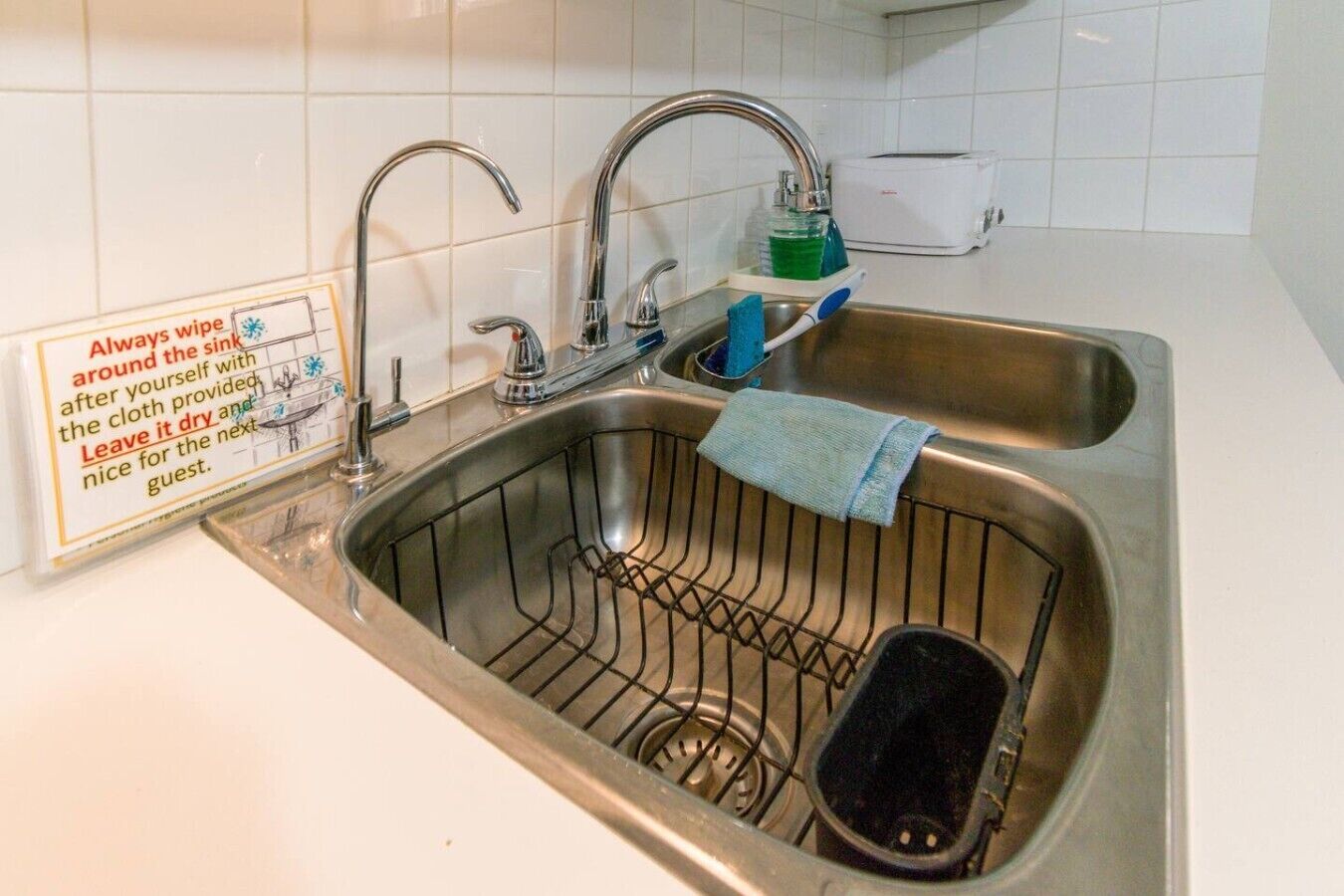 A stainless steel sink with a dish rack, a towel, and a soap dispenser. A sign instructs to always wipe the sink around with the cloth provided and leave it dry for the next guest.