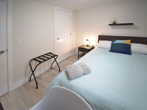 Ein Schlafzimmer mit Bett, Nachttisch mit Lampe, Handtuch auf dem Bett, Stuhl und einer Tür.