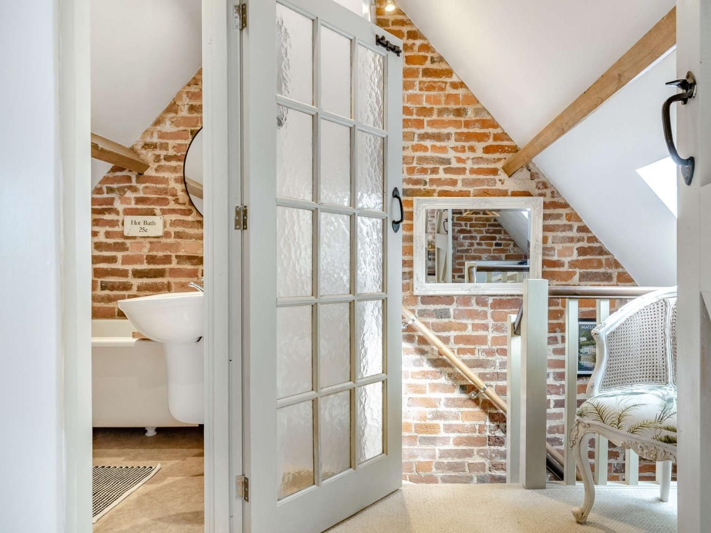 Una habitación con una bañera blanca, una puerta de vidrio, una pared de ladrillo y una silla blanca.