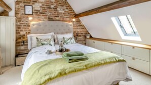 Un dormitorio con techo inclinado, paredes de ladrillo visto, cama con ropa de cama blanca, tragaluz y armario blanco.