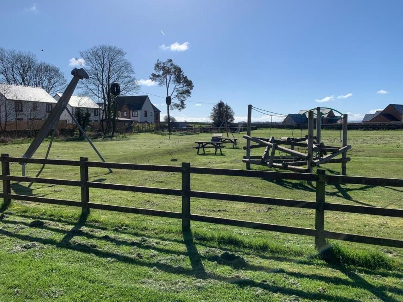 Un campo cubierto de césped con una valla de madera, mesas de picnic y un columpio.