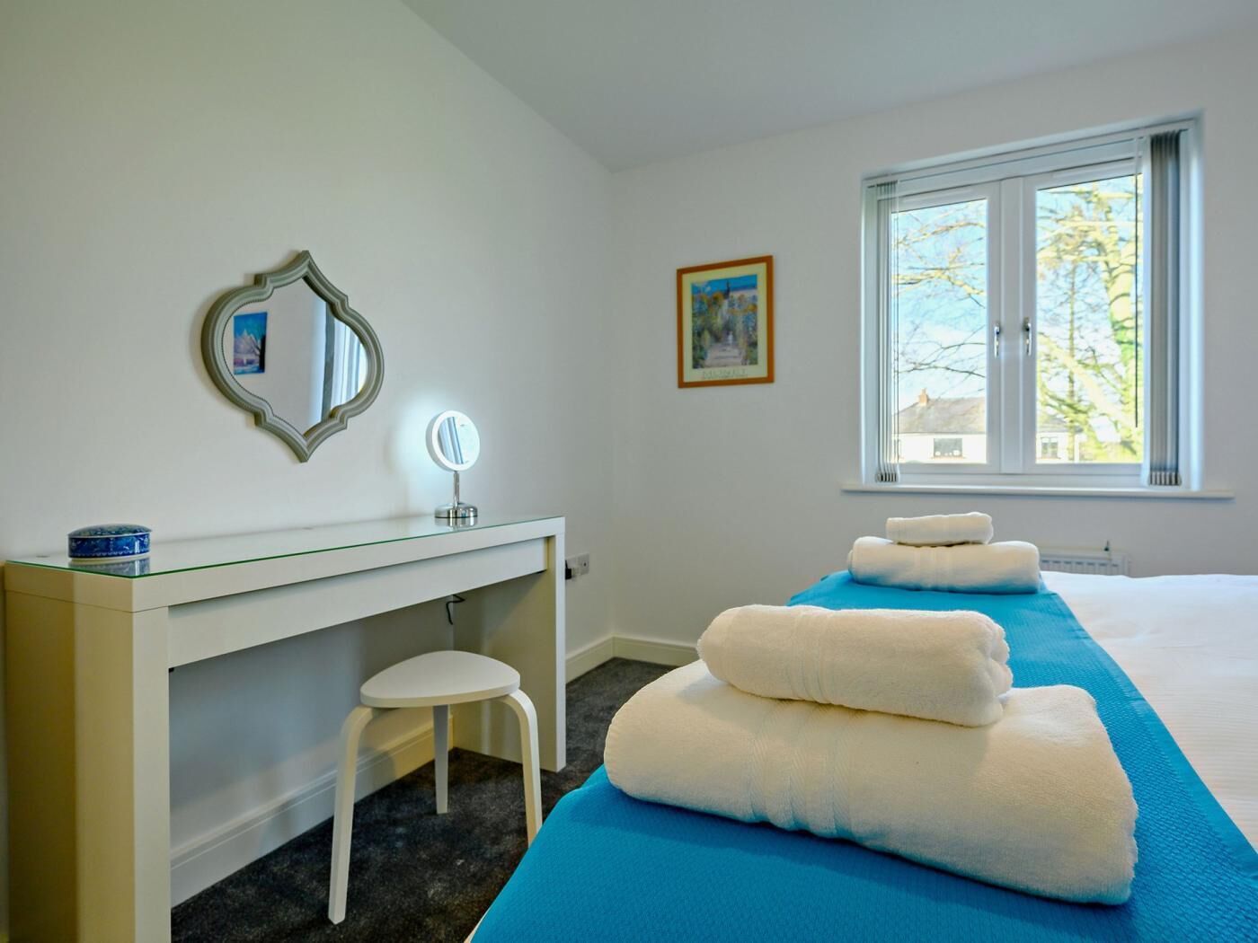 Habitación de hotel con cama, un escritorio con silla, espejo, ventana con vistas y un cuadro en la pared.