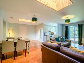 A modern living room with a brown leather sofa, a dining table with chairs, a kitchen area, and a large window with curtains. - 2 Bed in Sanquhar (oc-89403) (Sanquhar)