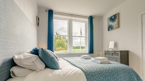 Een slaapkamer met een bed, een raam met gordijnen, een radiator, een bureau met lamp en een schilderij aan de muur.