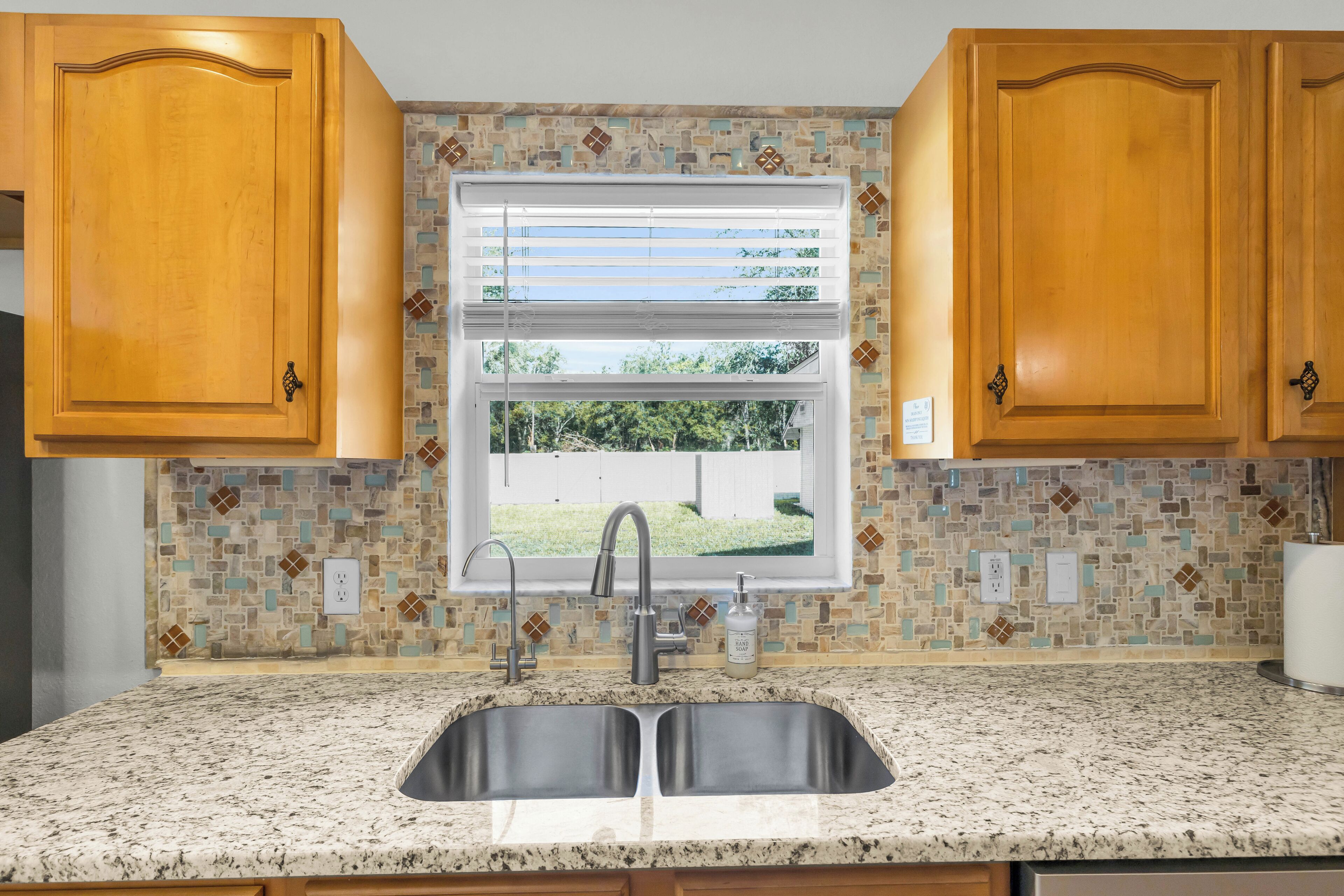A kitchen with wooden cabinets, a double sink, a window with blinds, and a mosaic backsplash.