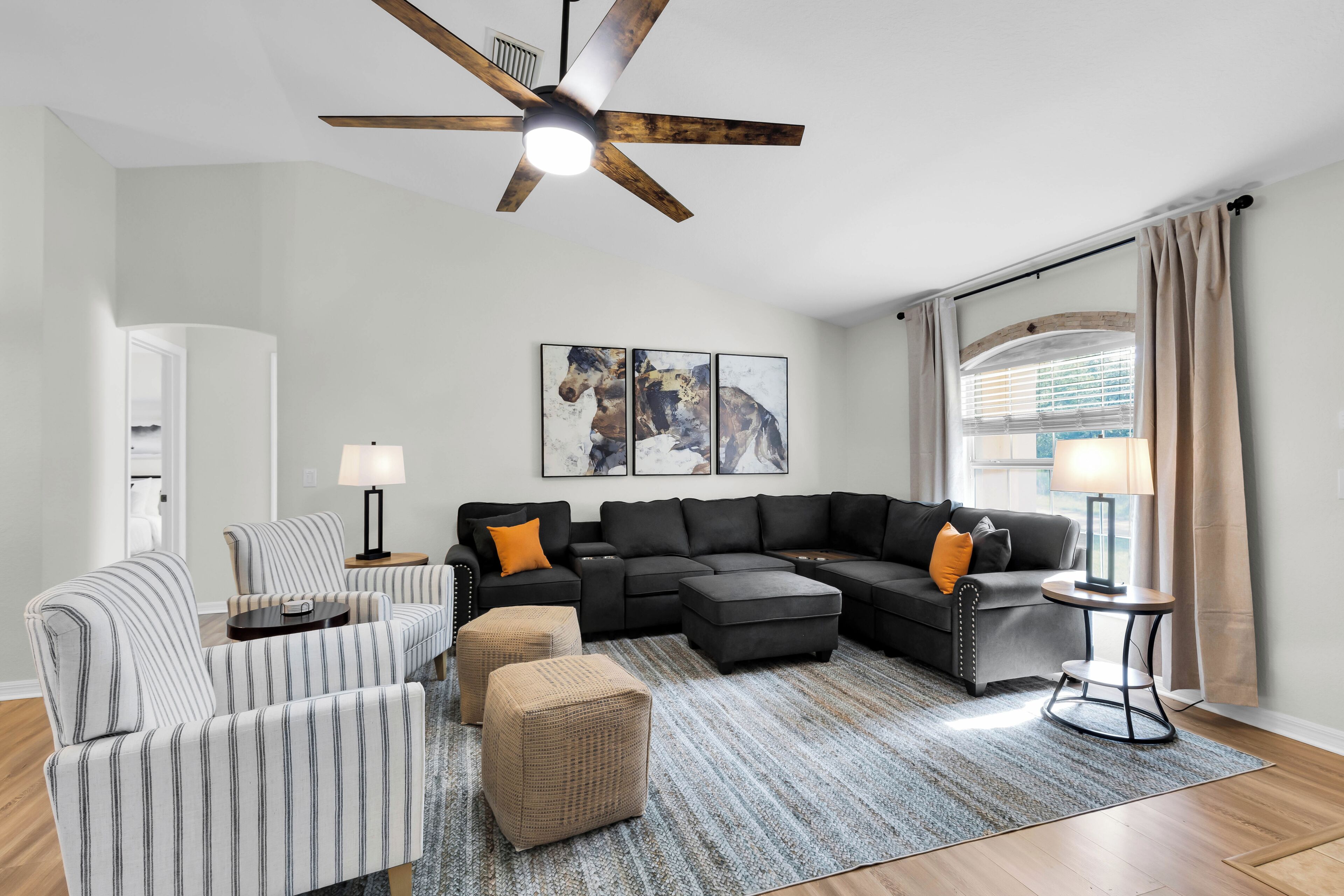 A living room with a sectional sofa, striped armchairs, a coffee table, a side table, a floor lamp, a ceiling fan, and artwork on the wall.