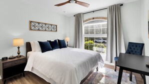 Une chambre d’hôtel comprenant un lit, des tables de chevet, une table, une chaise, un ventilateur de plafond, une fenêtre avec des rideaux et un élément décoratif mural.
