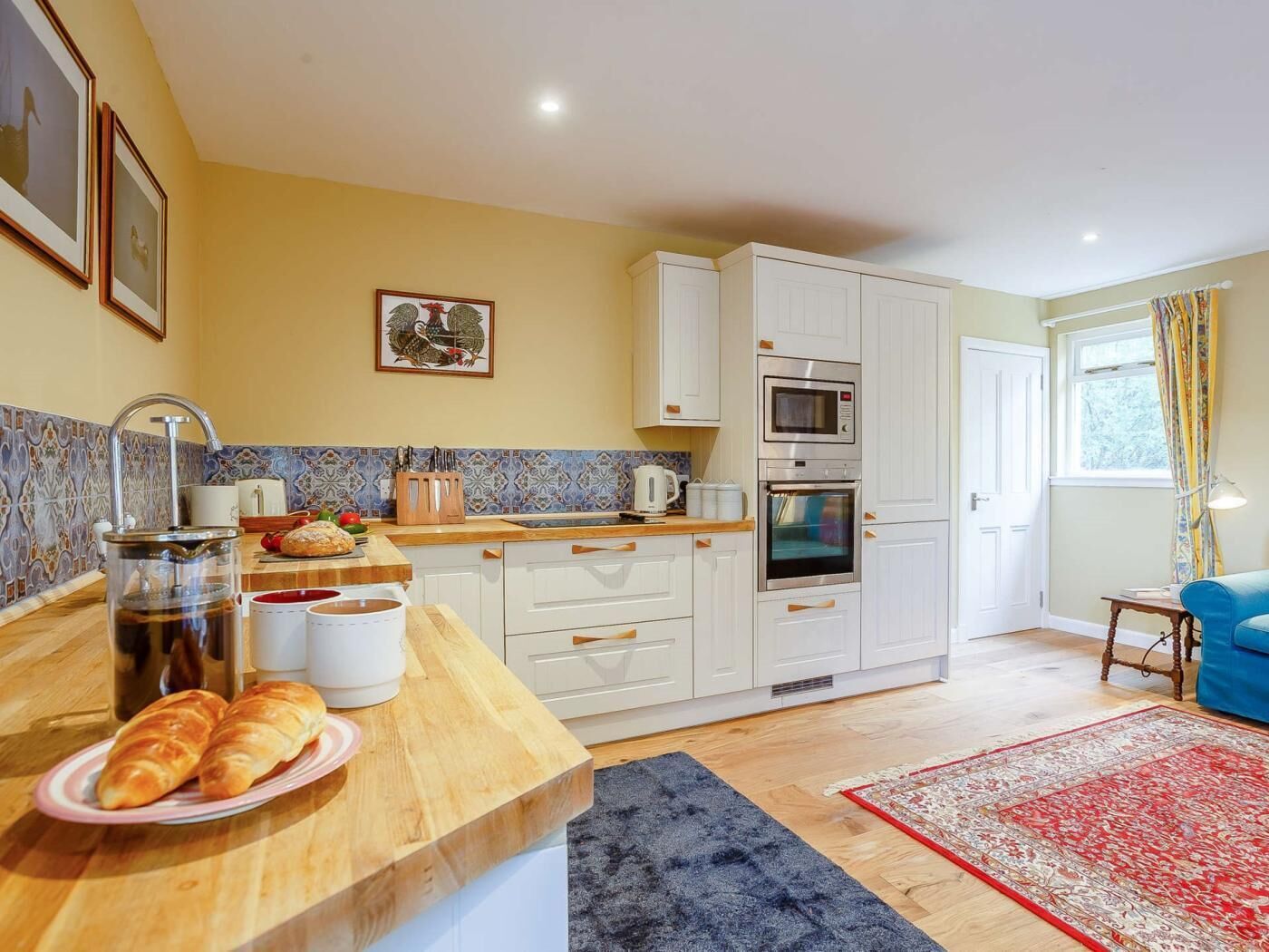 A modern kitchen with white cabinets, a microwave, oven, and a coffee maker. There is a wooden countertop with bread and a plate of croissants.