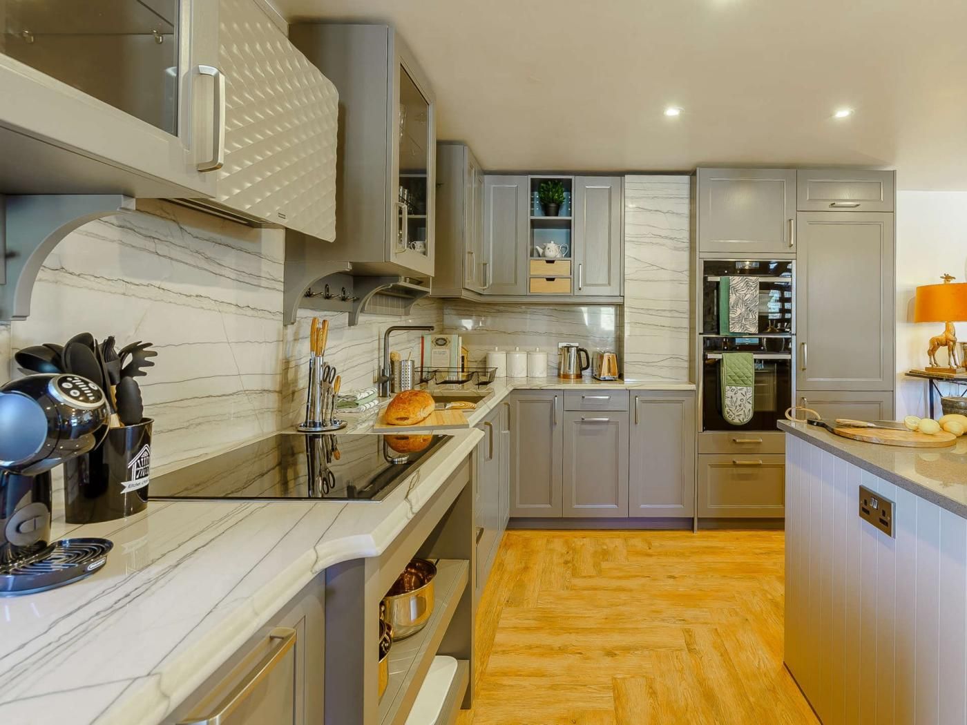 A modern kitchen with grey cabinets, a marble countertop, and a wooden herringbone floor.