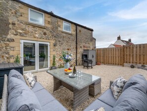 A stone house with a patio, outdoor seating, a barbecue, and a glass table with a vase of flowers. - 3 Bed in Berwick-upon-tweed (oc-89814) (Lowick)