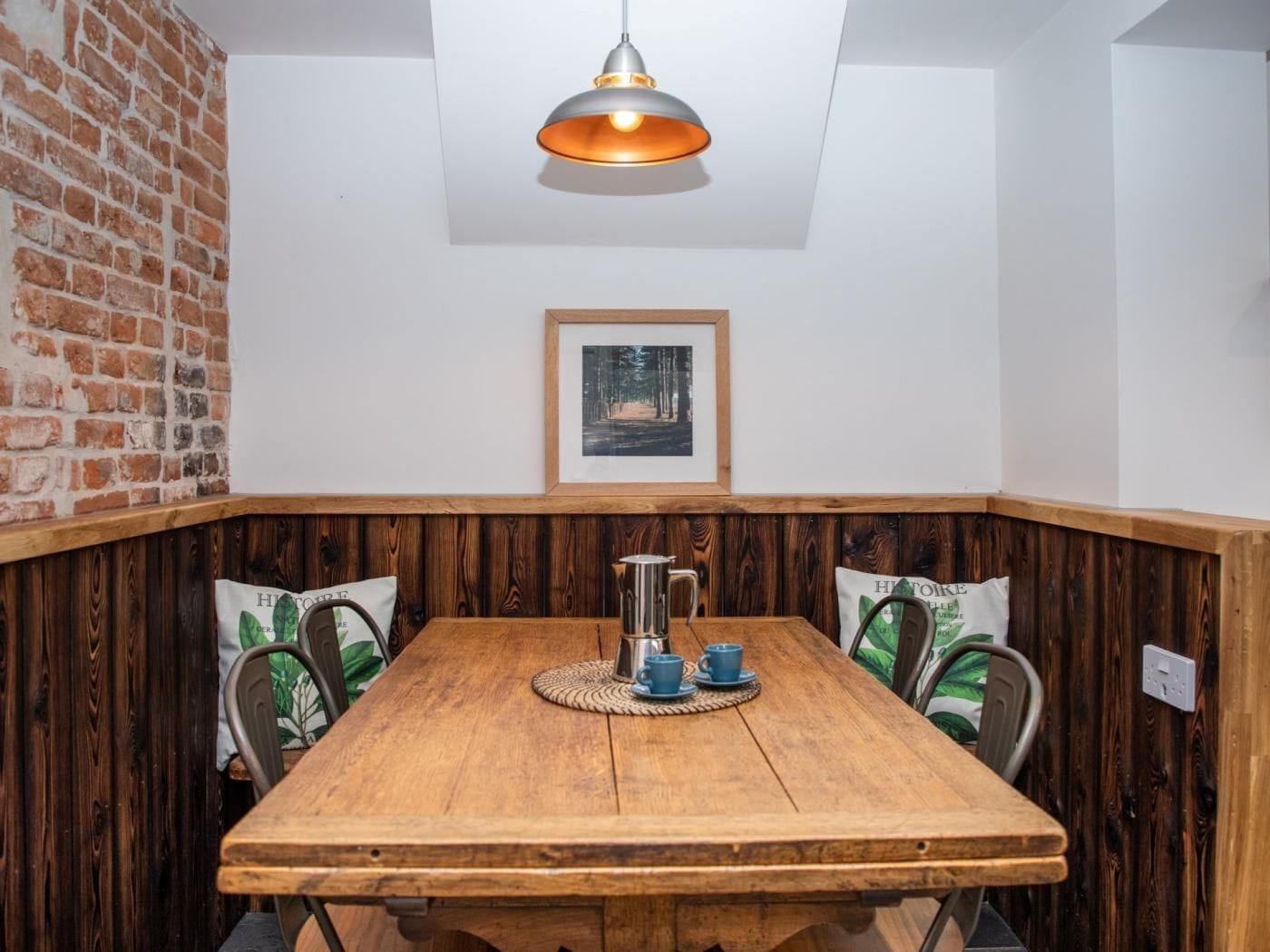Una mesa de comedor de madera con un jarro de metal y tazas azules, frente a una pared de ladrillo y paneles de madera.