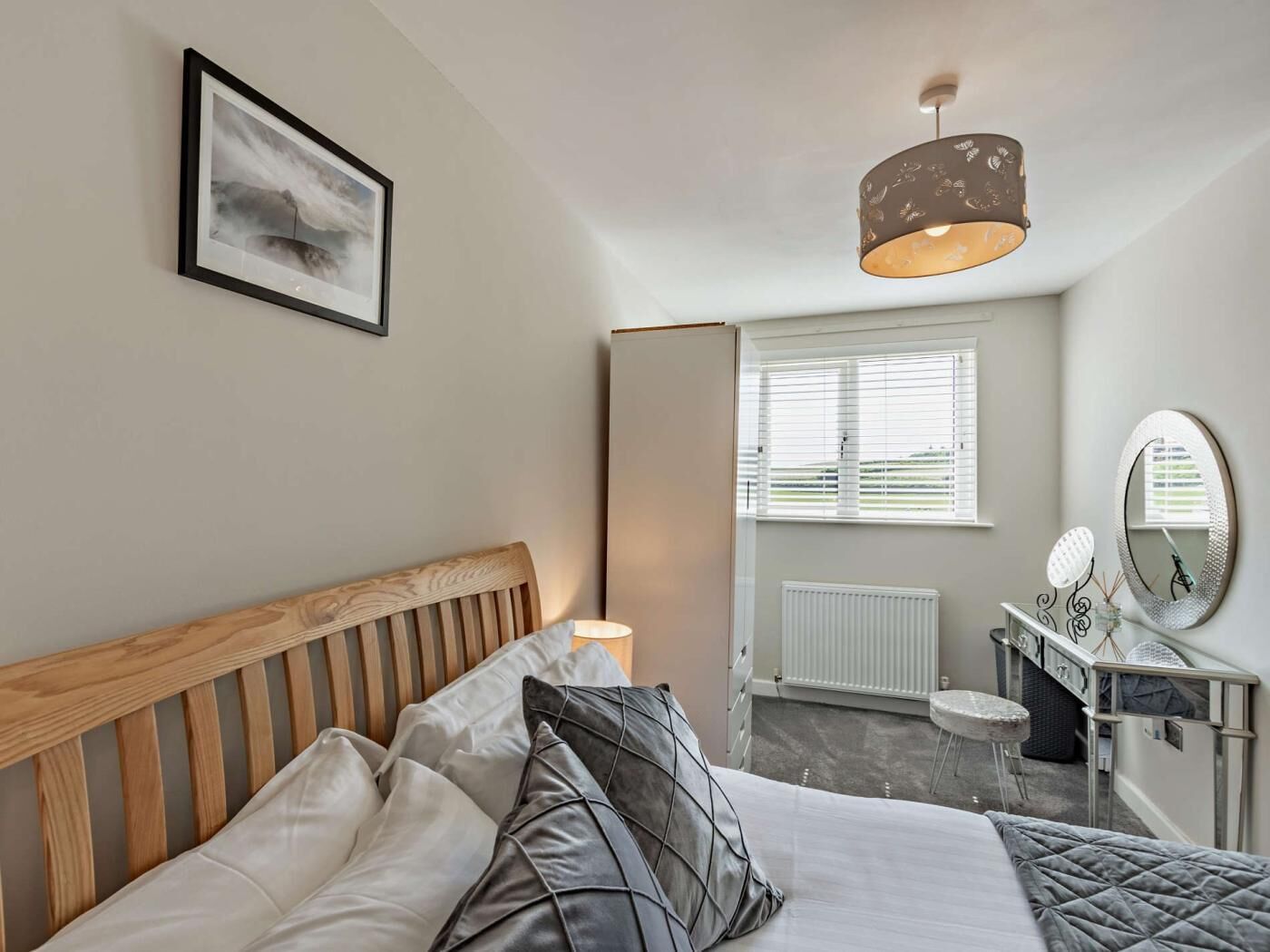 A bedroom with a wooden headboard, a bed with grey and black pillows, a bedside table with a mirror, and a window with blinds.