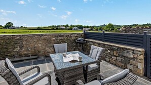 An outdoor patio with wicker furniture, a stone wall, and a view of a green field and houses. - 2 Bed in Grange-over-sands (oc-89206) (Cartmel)